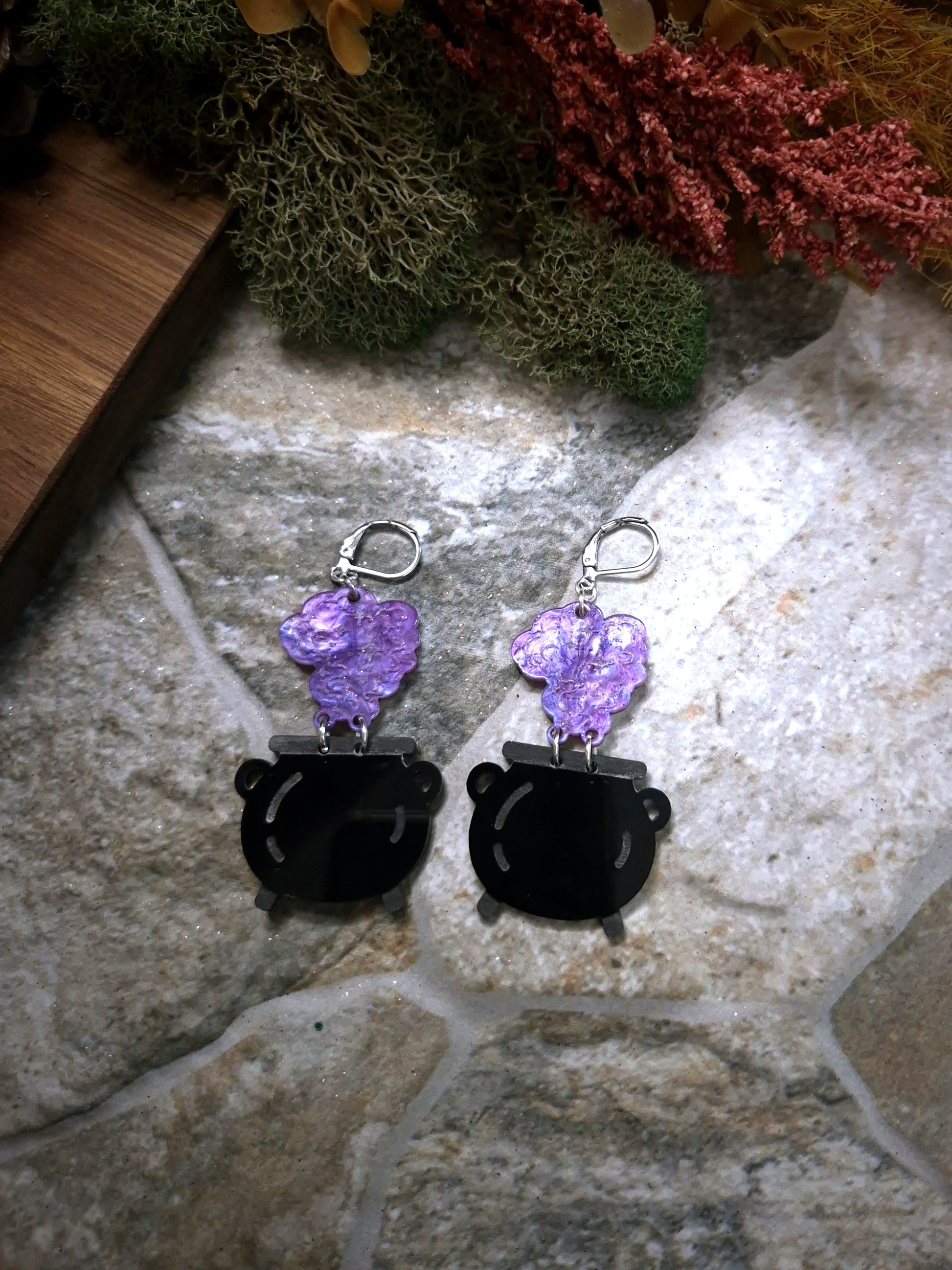 Cauldron earrings on a stone and wood background surrounded by foliage 
