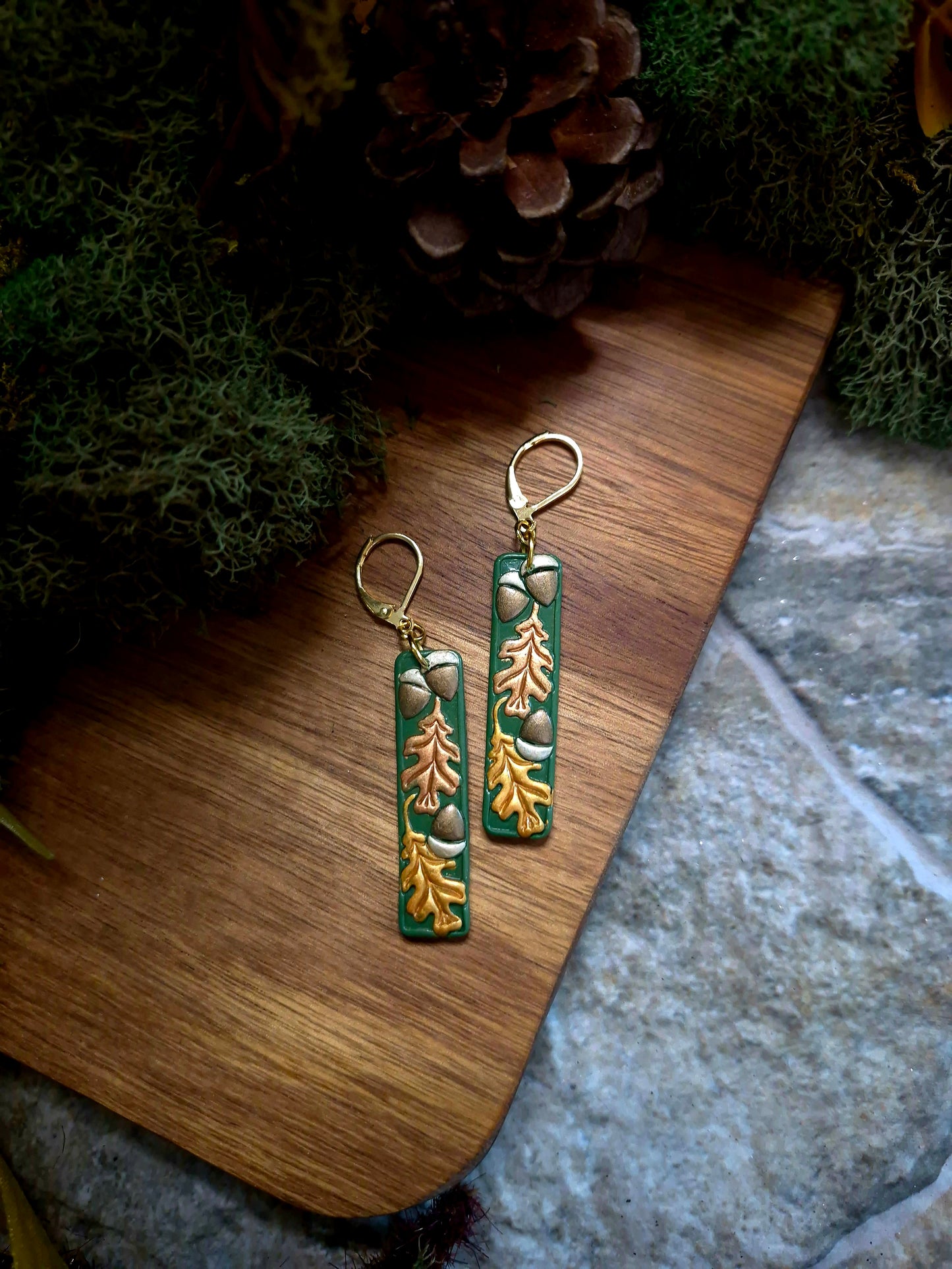 Green bar shaped earrings with metallic acorns and oak leaves on a stone and wood background surrounded by foliage. 