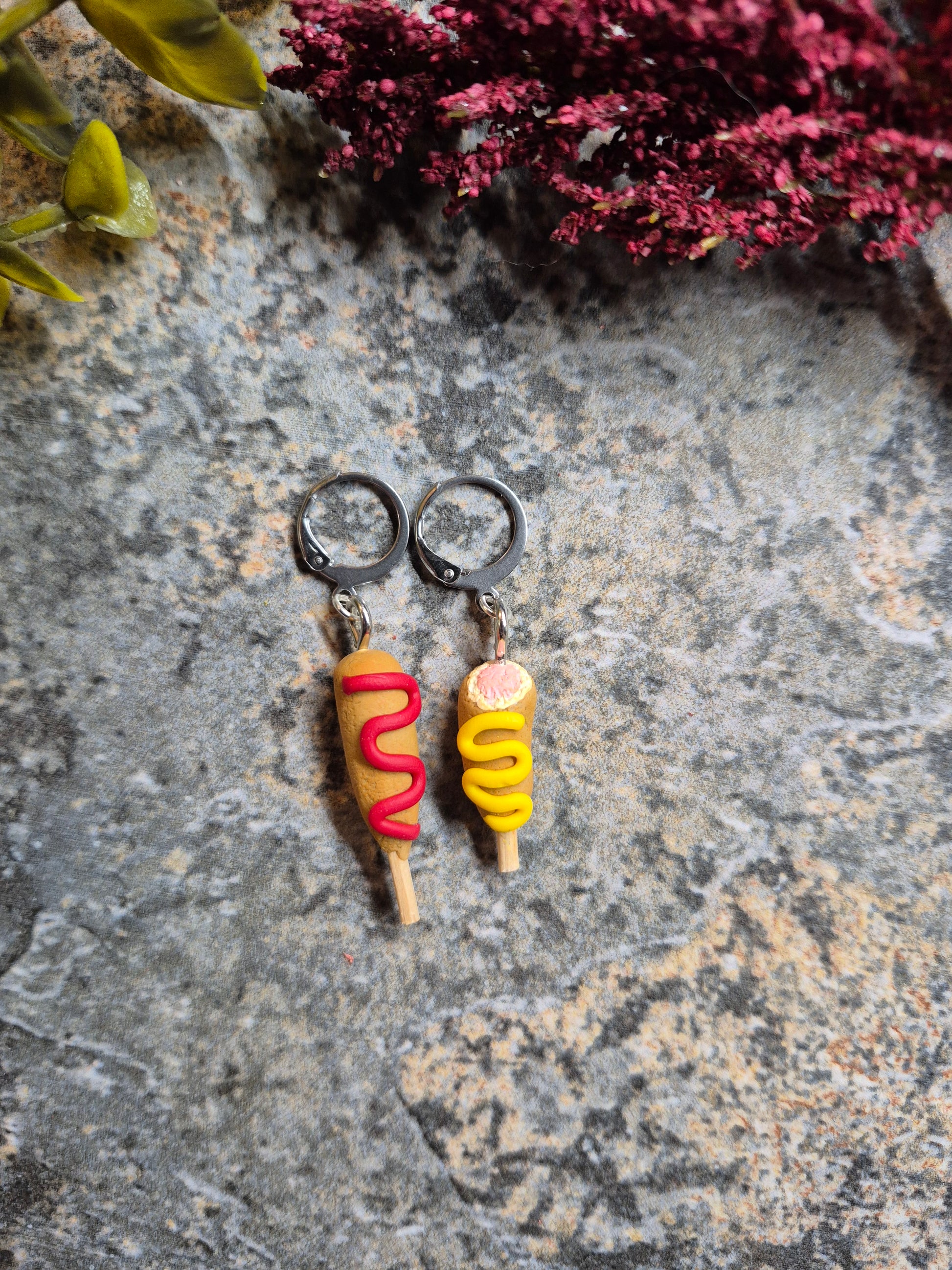 Full Corn Dog and Bitten Corn Dog Earrings on a stone background surrounded by foliage 
