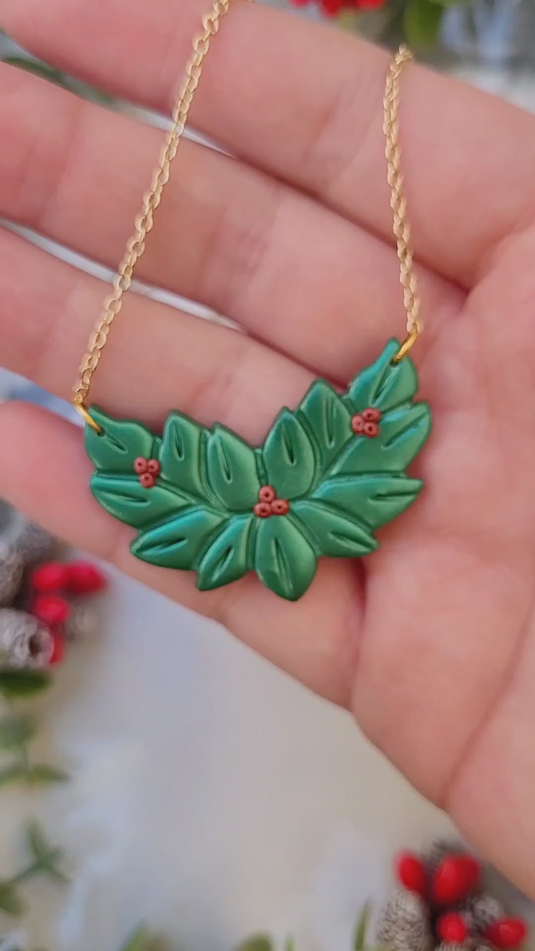 video close up of Metallic green and red holly wreath necklace on a marble background surrounded by foliage.