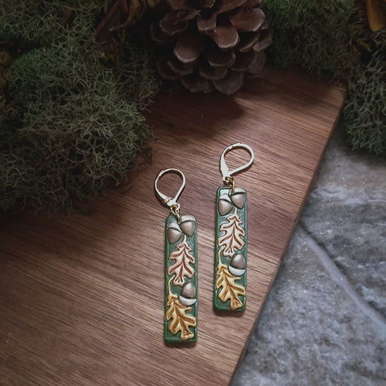 video close up of Green bar shaped earrings with metallic acorns and oak leaves on a stone and wood background surrounded by foliage. 