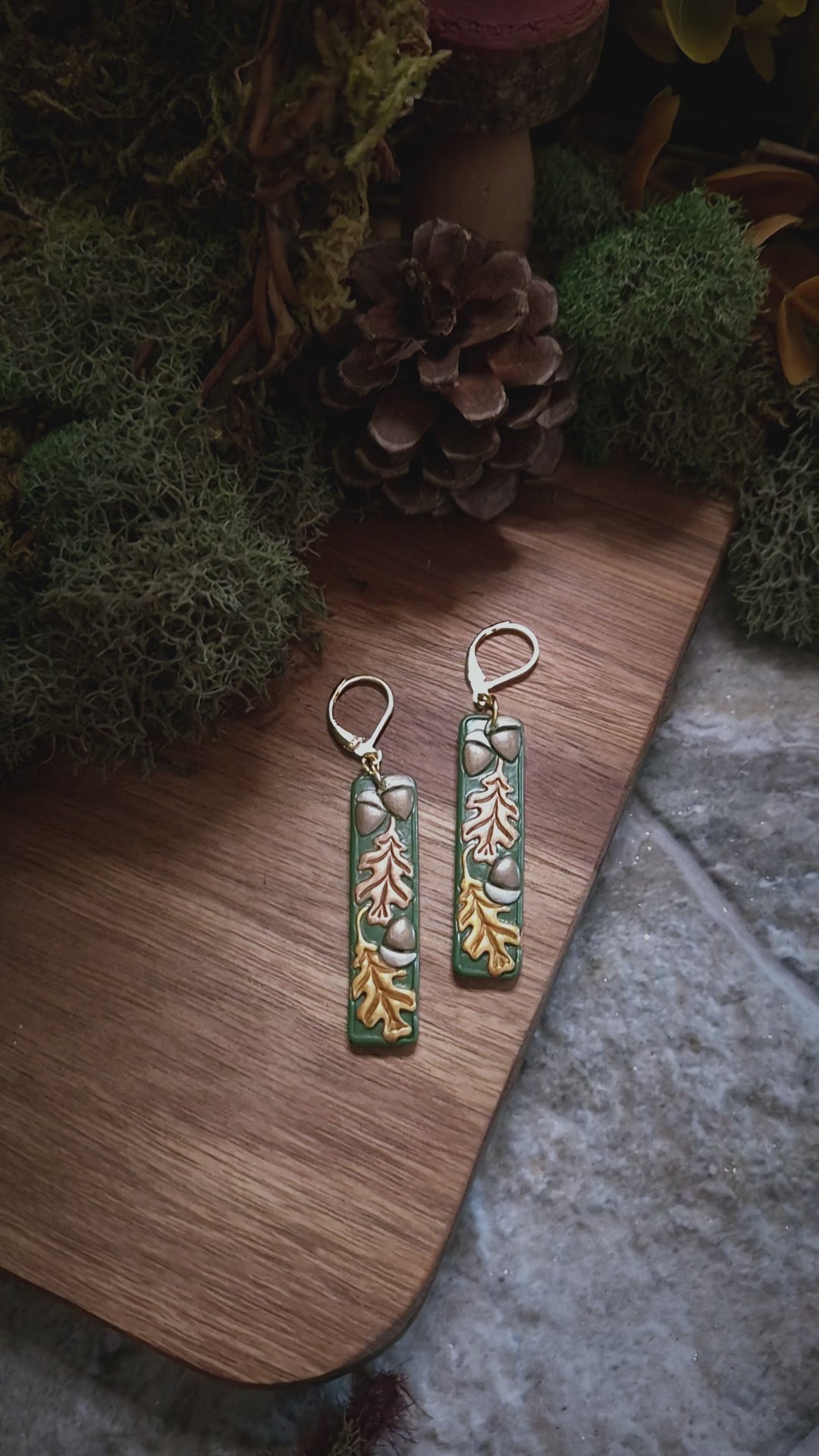 video close up of Green bar shaped earrings with metallic acorns and oak leaves on a stone and wood background surrounded by foliage. 