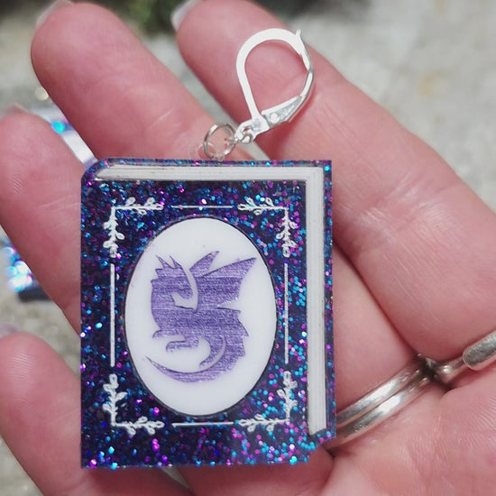 video of Glitter Dragon Book Earrings on a stone background surrounded by foliage. 