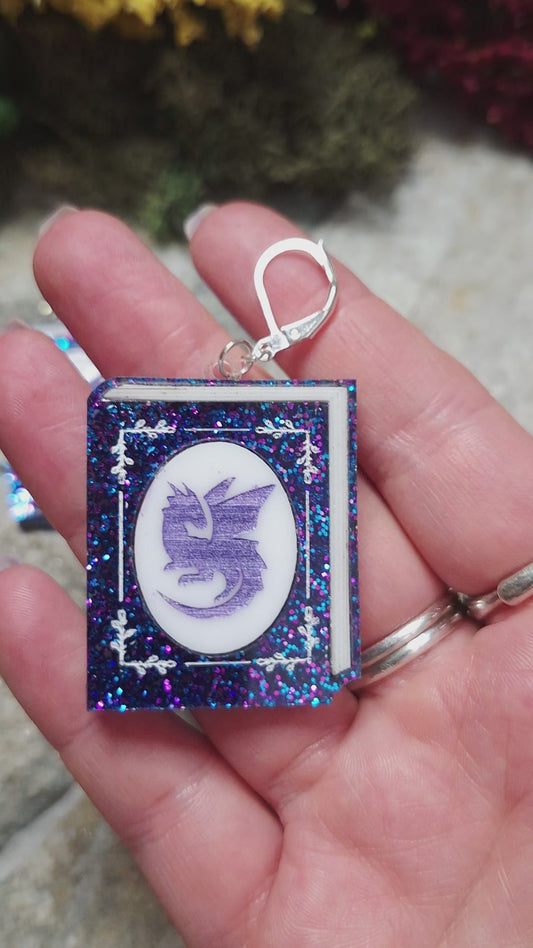 video of Glitter Dragon Book Earrings on a stone background surrounded by foliage. 