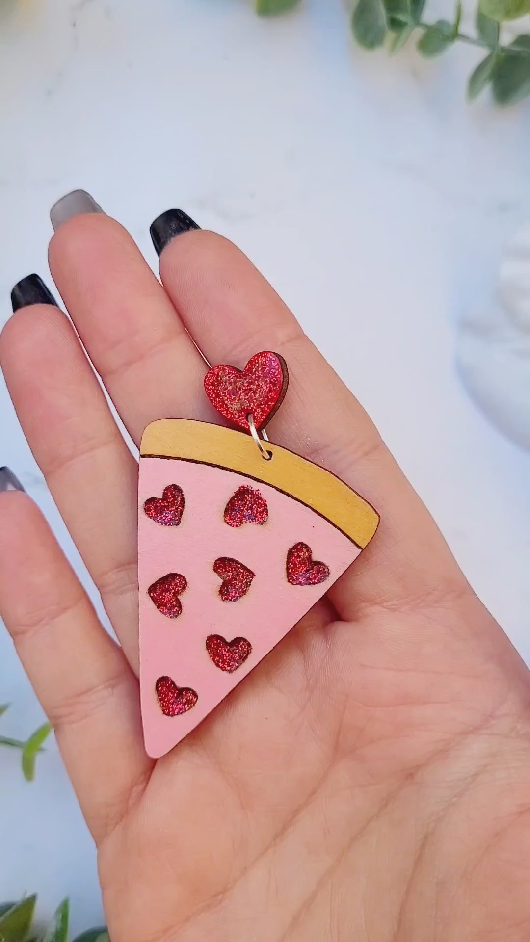 video close up of slice of love pizza earrings on a white marble background surrounded by foliage.