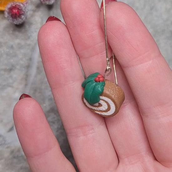 video close up of yule log earrings on a wood and stone background surrounded by foliage. 