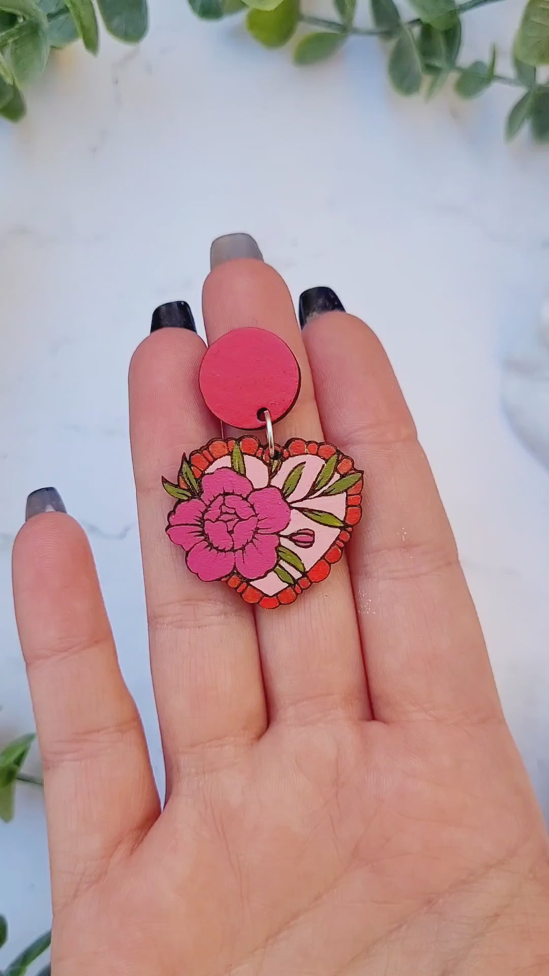 video close up of Rose tattoo earrings on a white marble background surrounded by foliage.