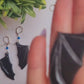video close up of bat boy earrings on a marble background surrounded by foliage.
