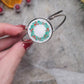 Video close up of silver holly bracelet on a wood and stone background surrounded by foliage. 