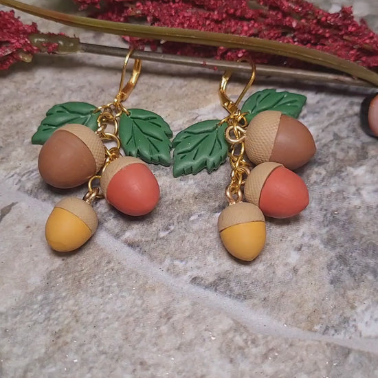 video close up of 3d acorn shaped earrings with  oak leaves. Sitting on a stone background surrounded by foliage. 