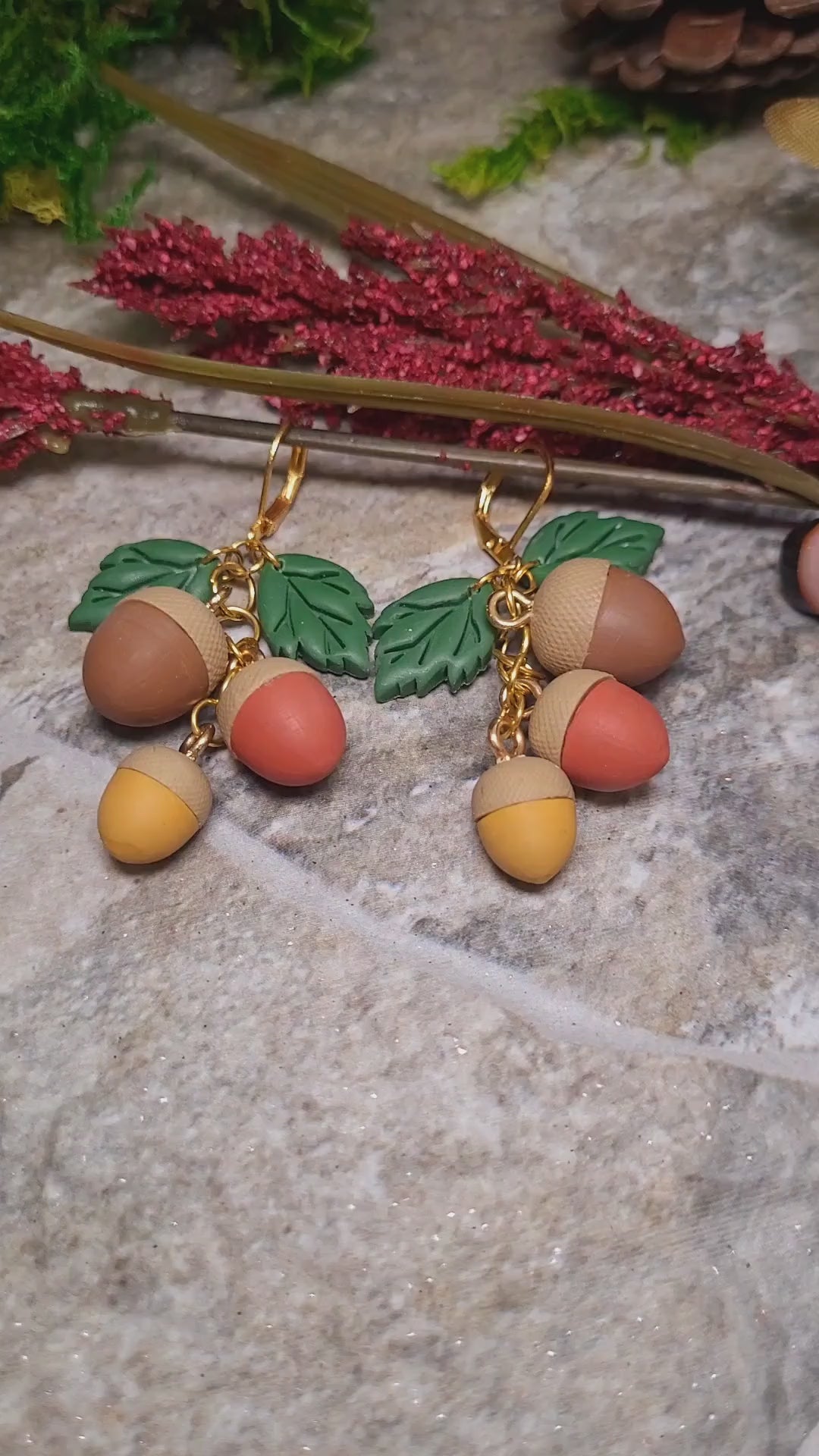 video close up of 3d acorn shaped earrings with  oak leaves. Sitting on a stone background surrounded by foliage. 