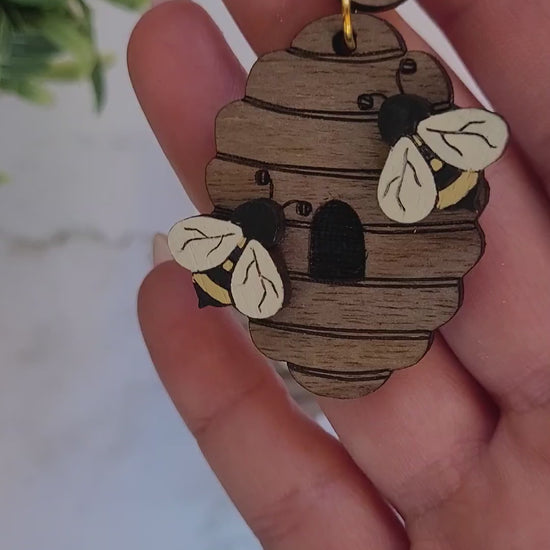 video close up of wooden beehive shaped earrings with bee charms on a marble background surrounded by foliage.