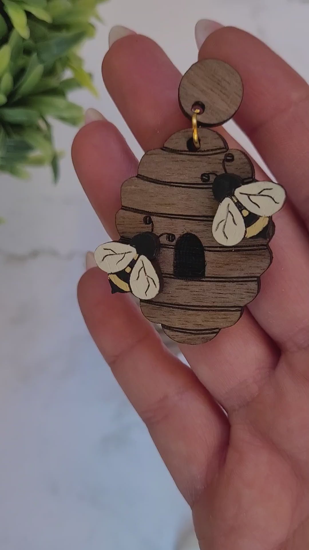 video close up of wooden beehive shaped earrings with bee charms on a marble background surrounded by foliage.