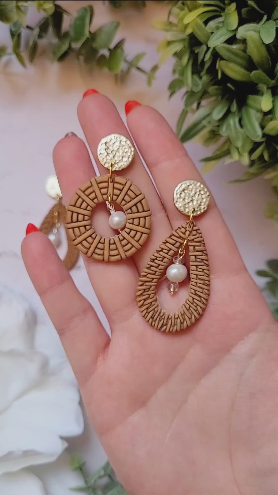 video close up of tear drop and round rattan earrings on a marble background with foliage.