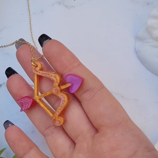 video close up of gold and pink cupid's bow necklace on a white marble background surrounded by foliage.