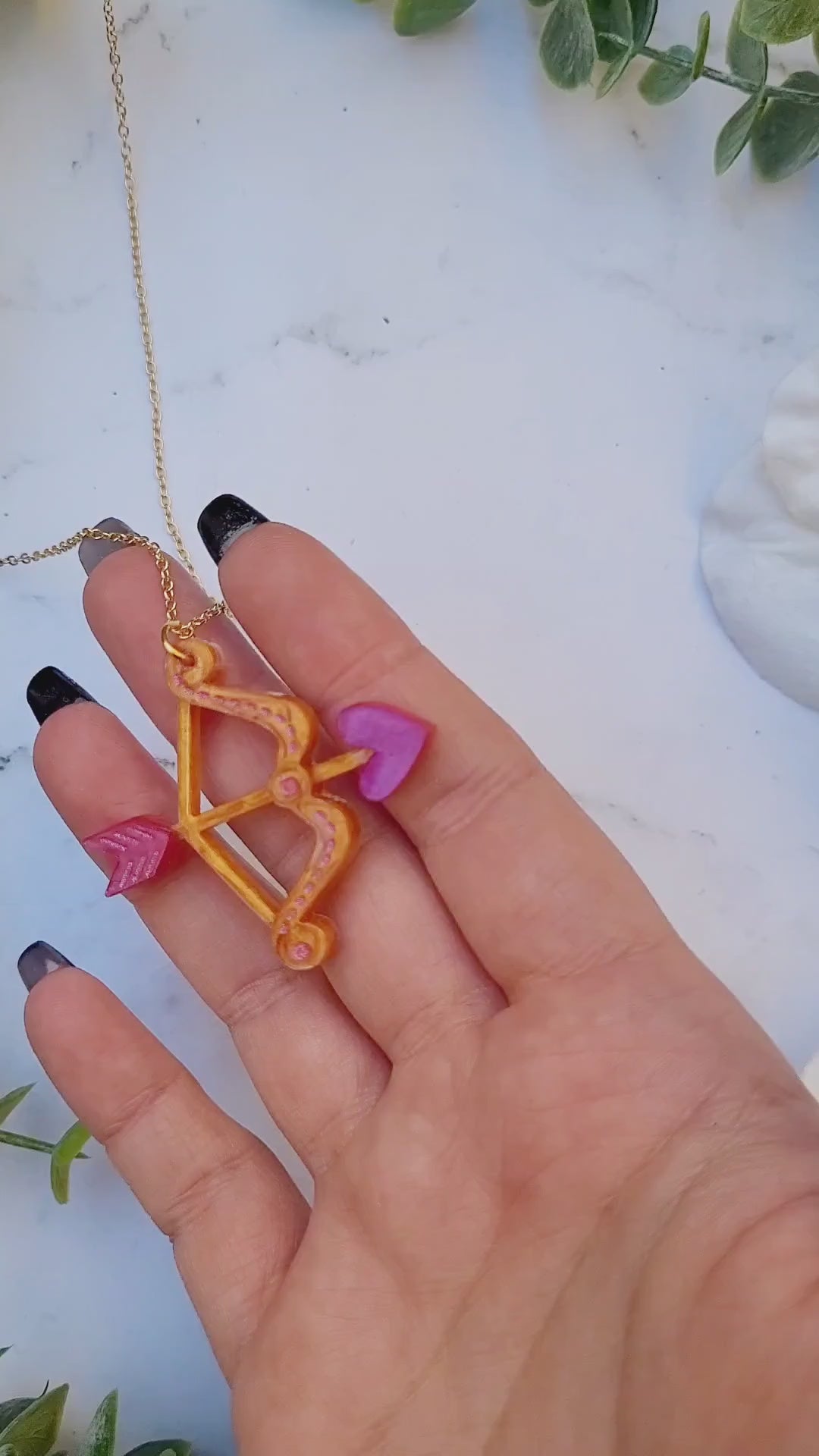 video close up of gold and pink cupid's bow necklace on a white marble background surrounded by foliage.
