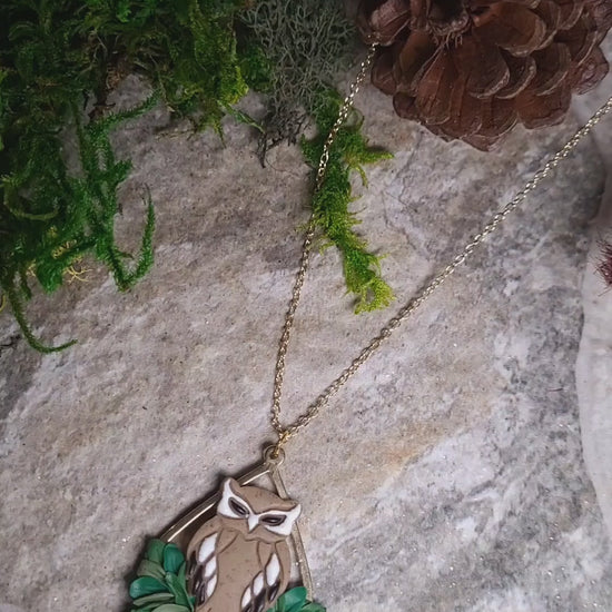 video close up of Owl necklace sitting on a stone background surrounded by foliage. 