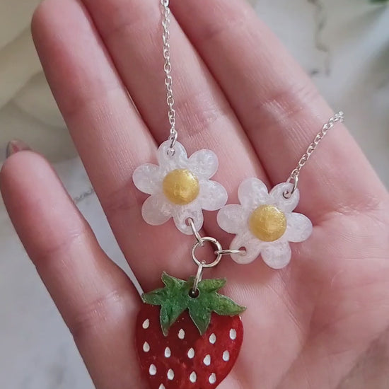 video close up of Resin Strawberry and flower necklace on a marble background surrounded by foliage.