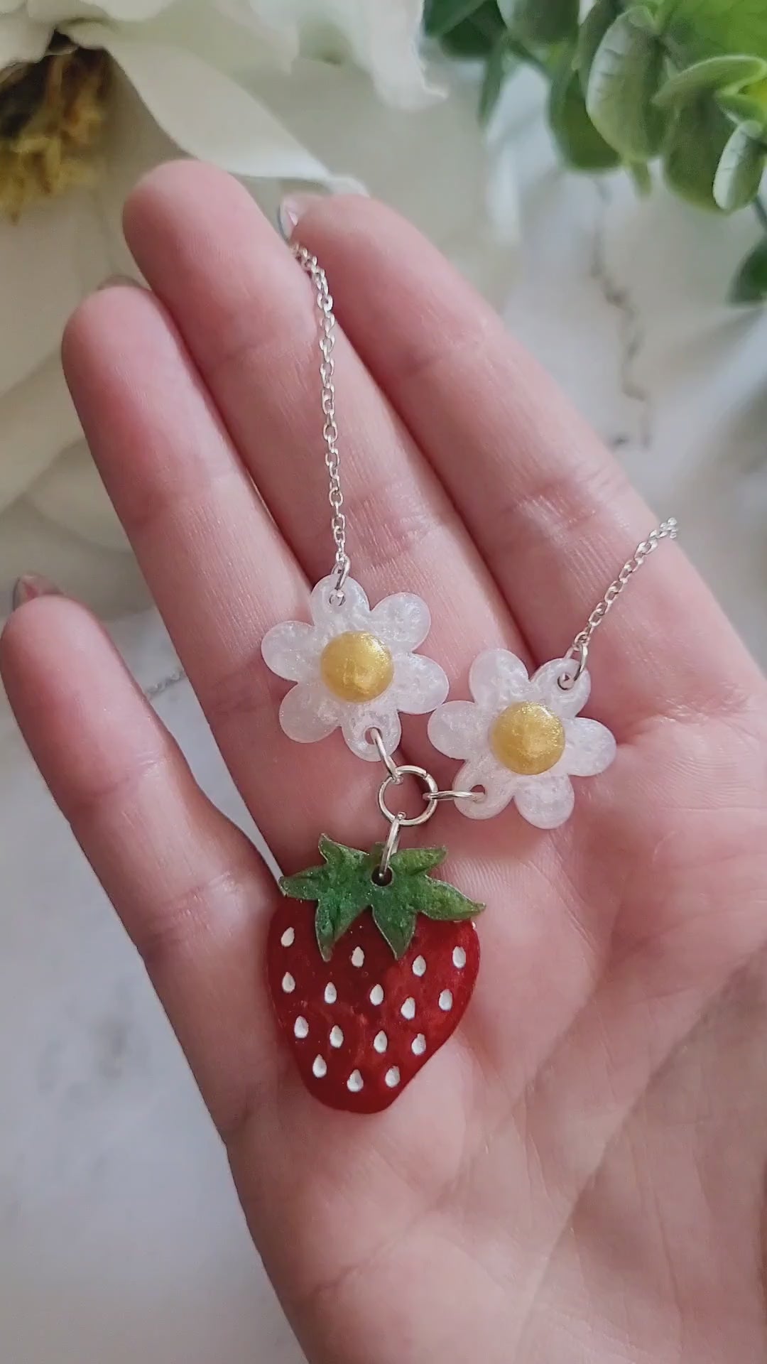 video close up of Resin Strawberry and flower necklace on a marble background surrounded by foliage.
