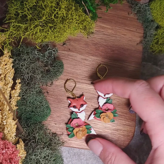 video close up of floral fox earrings on wood background surrounded by foliage. 