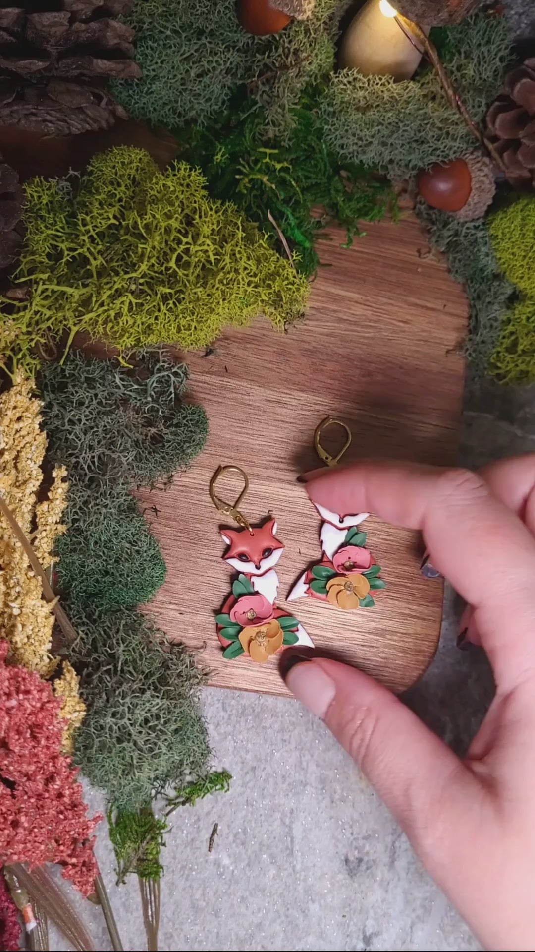 video close up of floral fox earrings on wood background surrounded by foliage. 