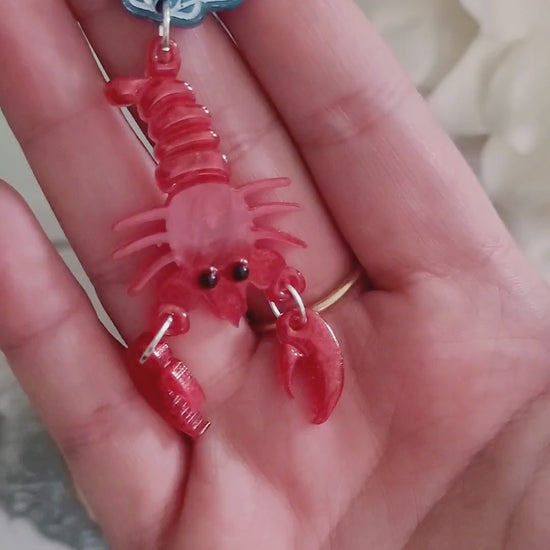 video close up of Red Lobster with a blue shell stud earrings on a marble background with foliage.
