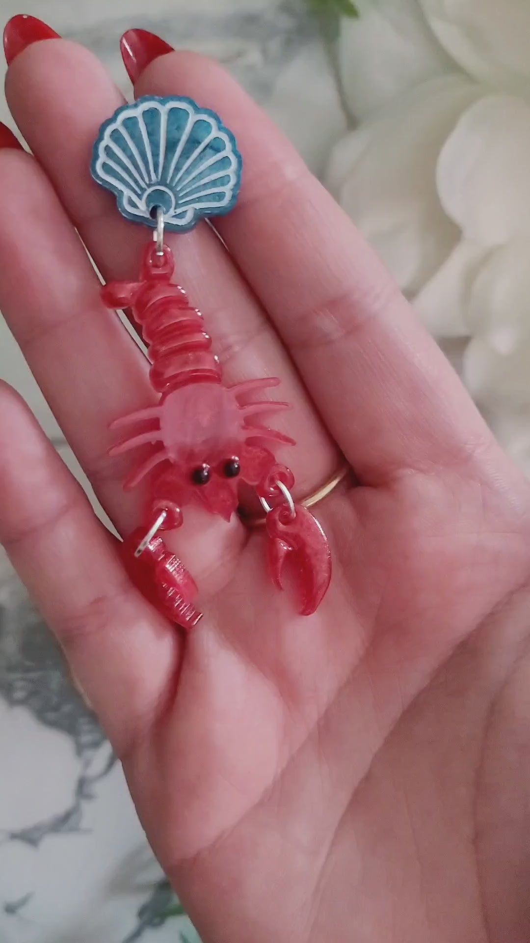 video close up of Red Lobster with a blue shell stud earrings on a marble background with foliage.