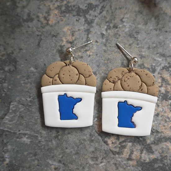 video of State fair cookie bucket earrings on a stone background surrounded by foliage .
