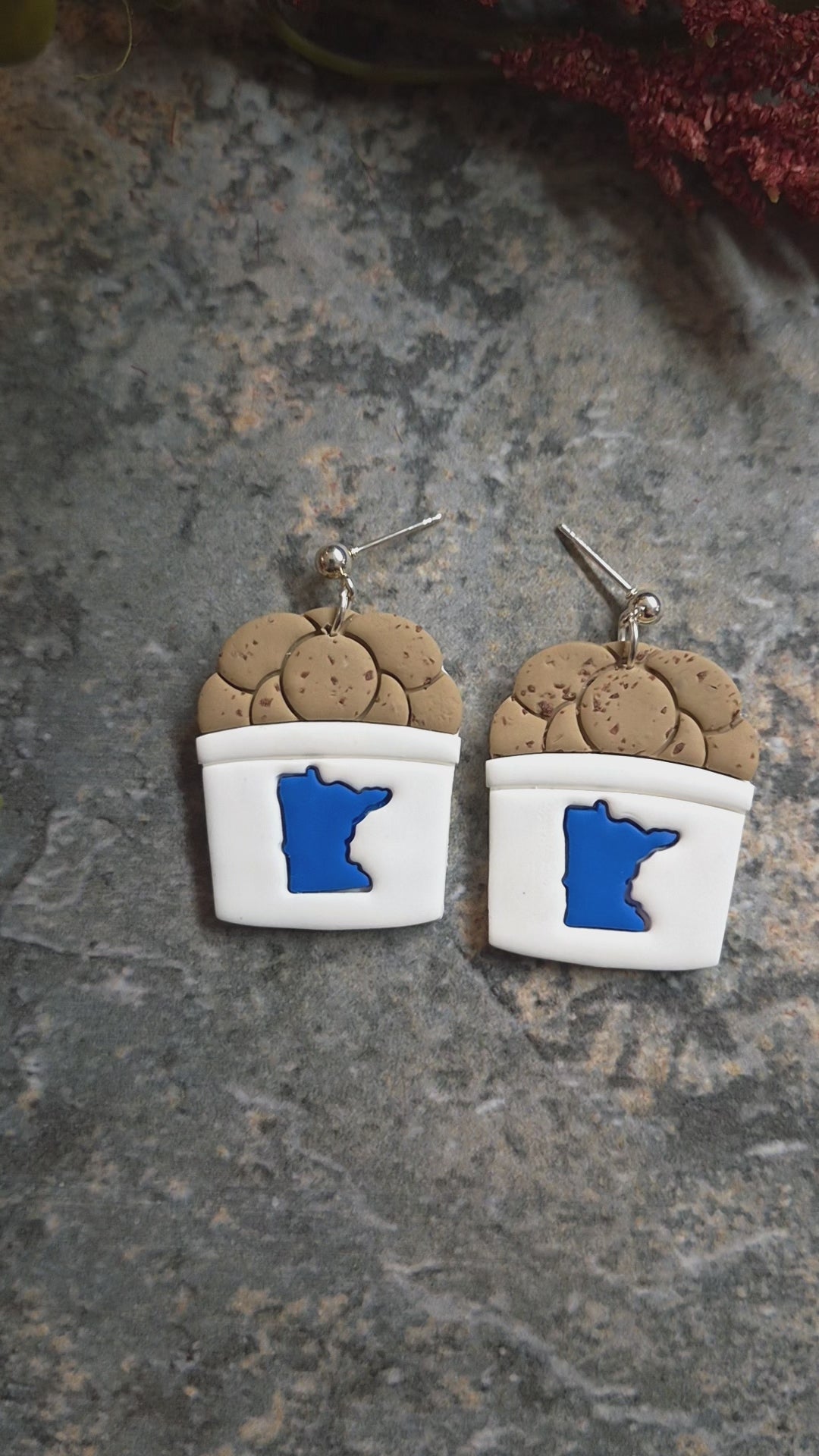 video of State fair cookie bucket earrings on a stone background surrounded by foliage .