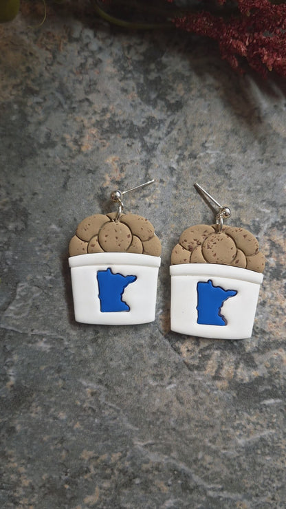 video of State fair cookie bucket earrings on a stone background surrounded by foliage .