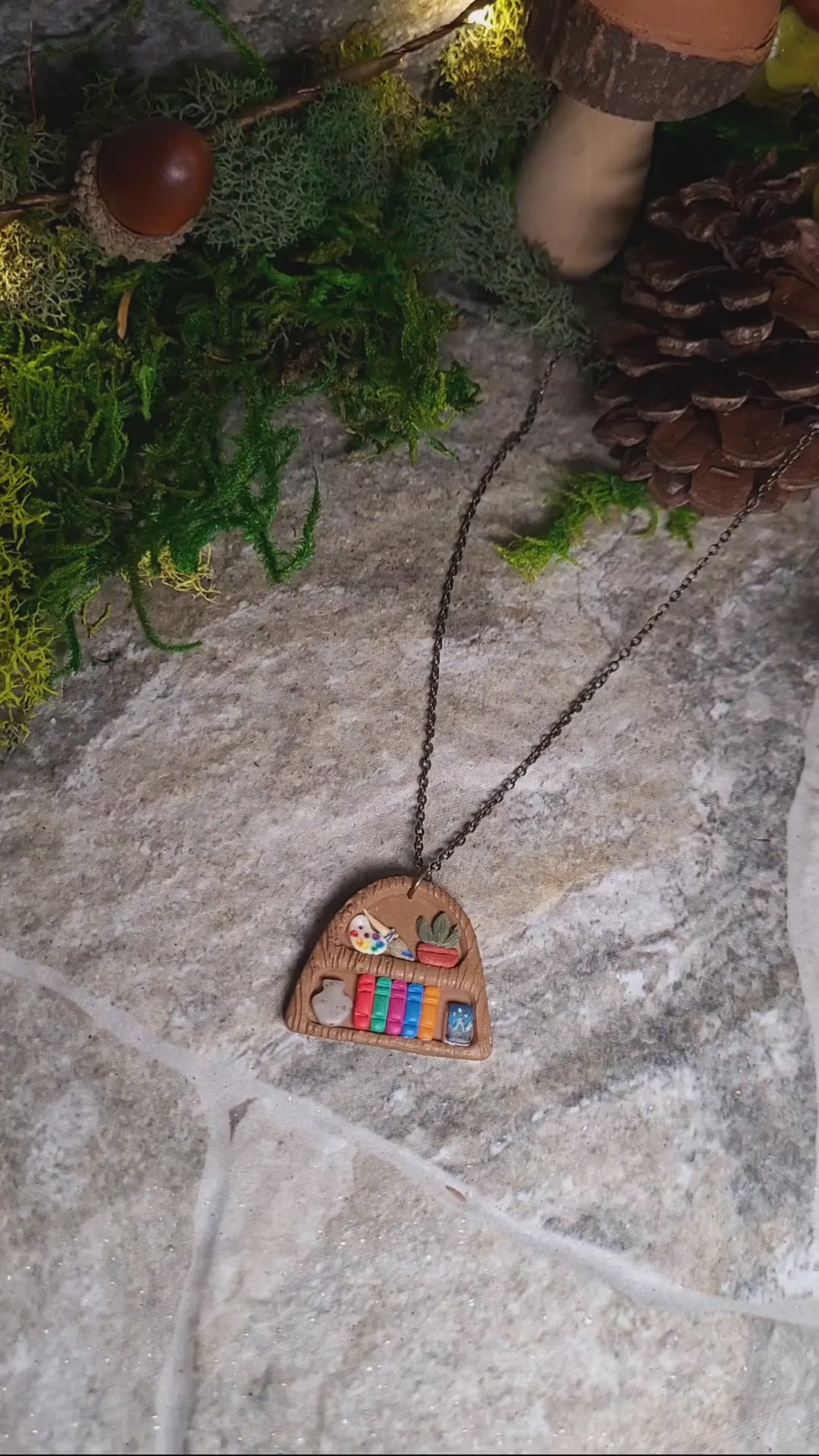 video close up of Bookshelf pendant made of polymer clay sits on a stone background surrounded by foliage. 