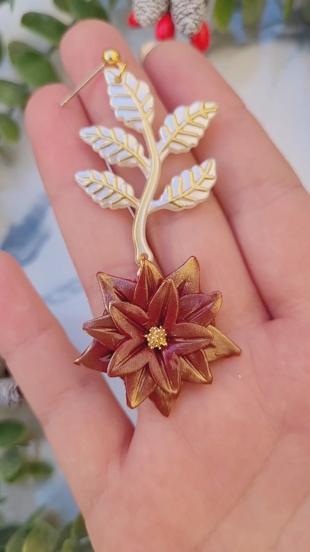 video close up of Red and gold poinsettia earrings on a marble background surrounded by foliage. 