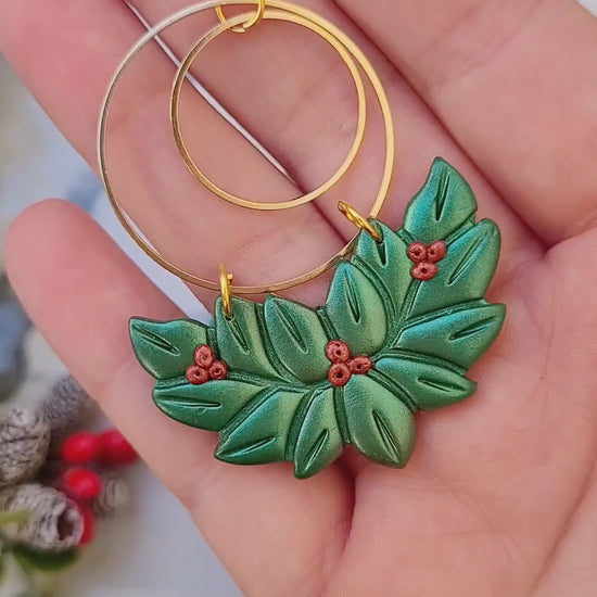video close up of Holly wreath earrings on a marble background surrounded by foliage.