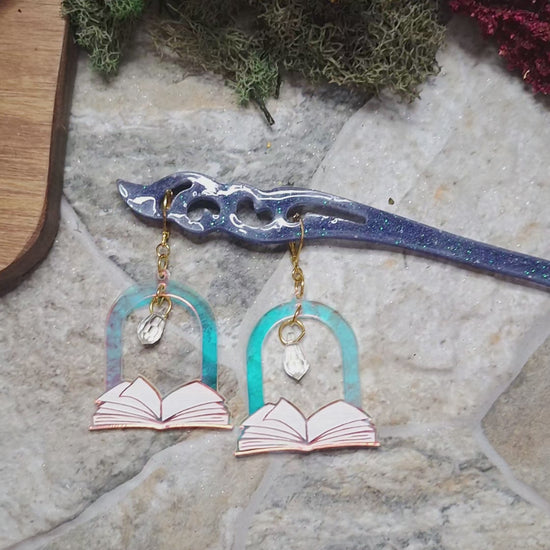 video of Open Book Arch earrings on a hair stick catching the light against a stone background surrounded by foliage 
