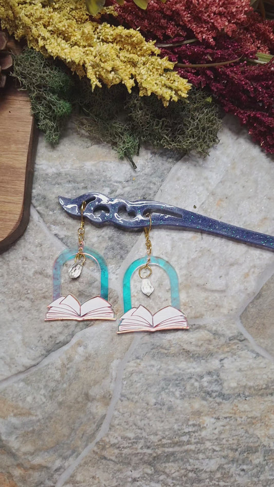 video of Open Book Arch earrings on a hair stick catching the light against a stone background surrounded by foliage 
