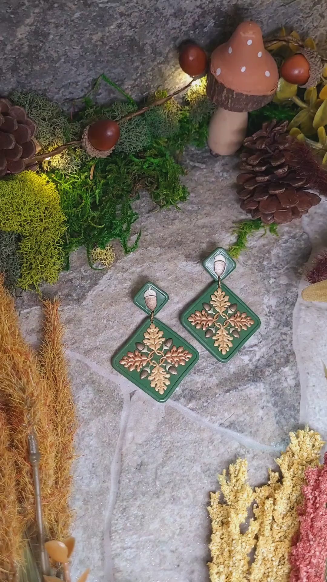 video close up of Green diamond shaped earrings with metallic acorns and oak leaves. Sitting on a stone background surrounded by foliage. 