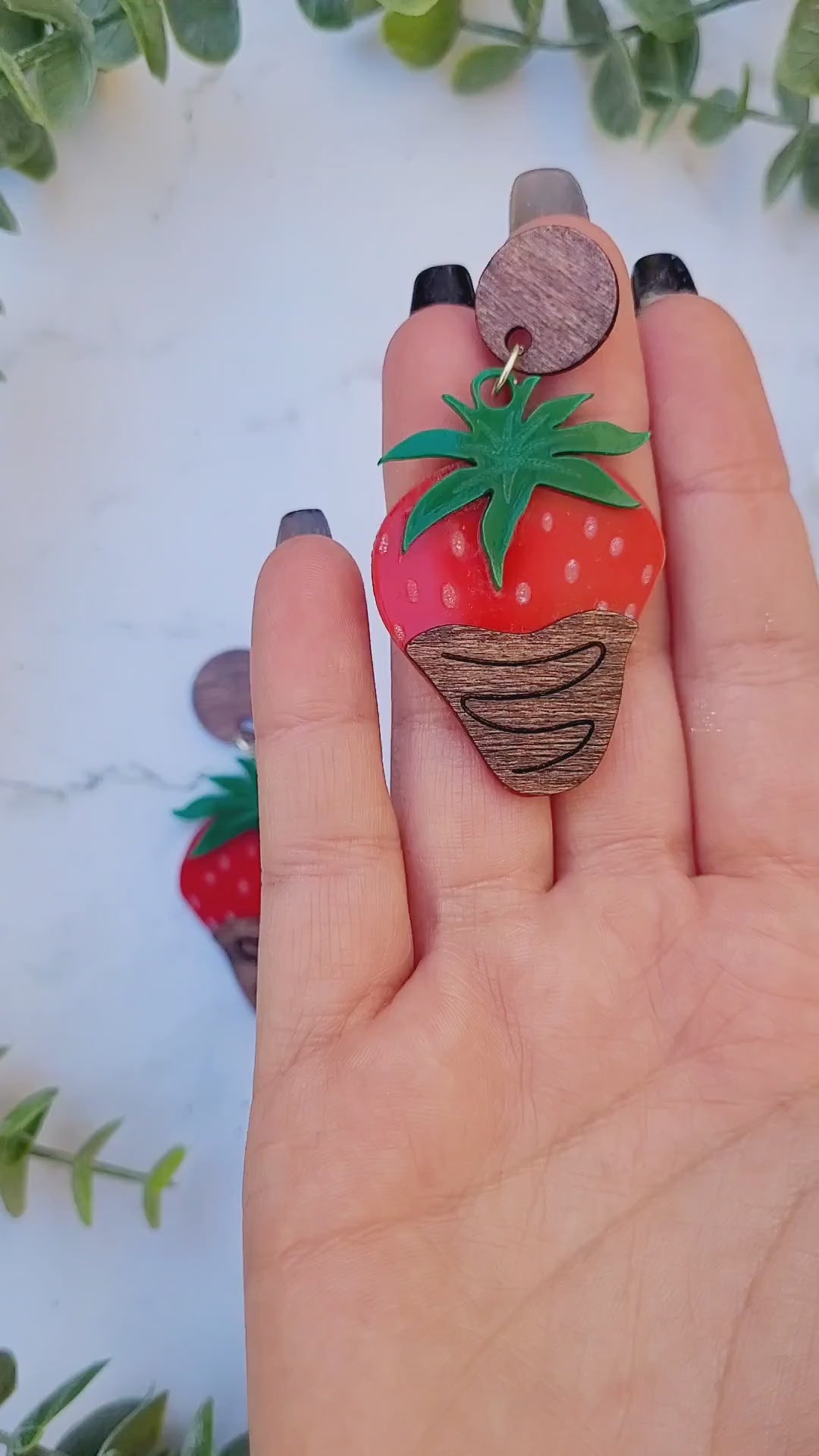 video close up of Chocolate covered strawberry earring on white marble background surrounded by foliage.
