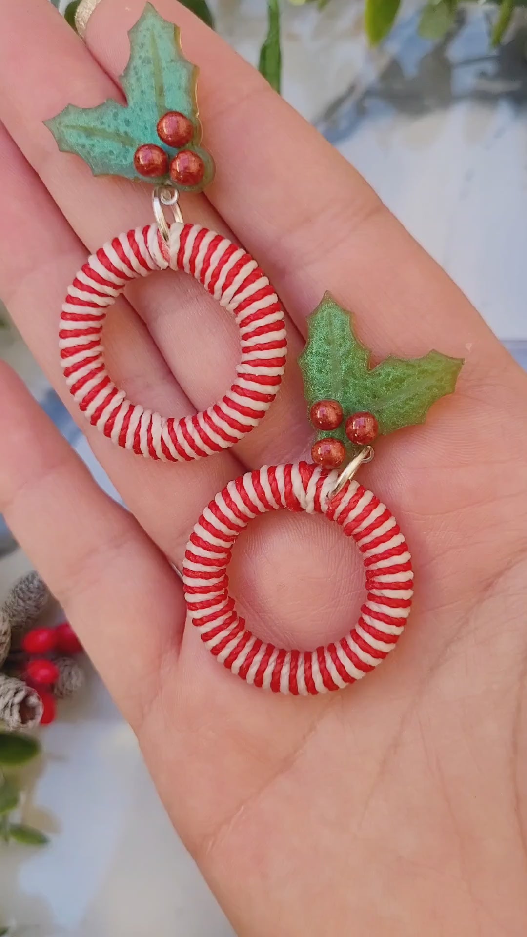 video close up of mistletoe and candy cane striped hoop earrings on a white marble background surrounded by foliage.