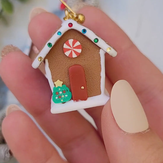 video close up of Gingerbread house earrings on a marble background surrounded by foliage.