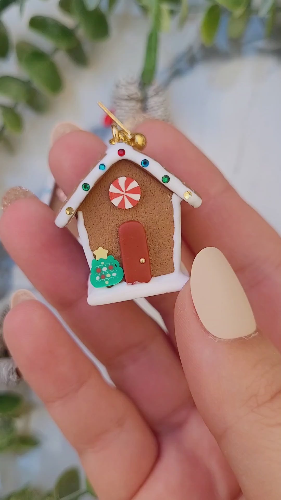 video close up of Gingerbread house earrings on a marble background surrounded by foliage.