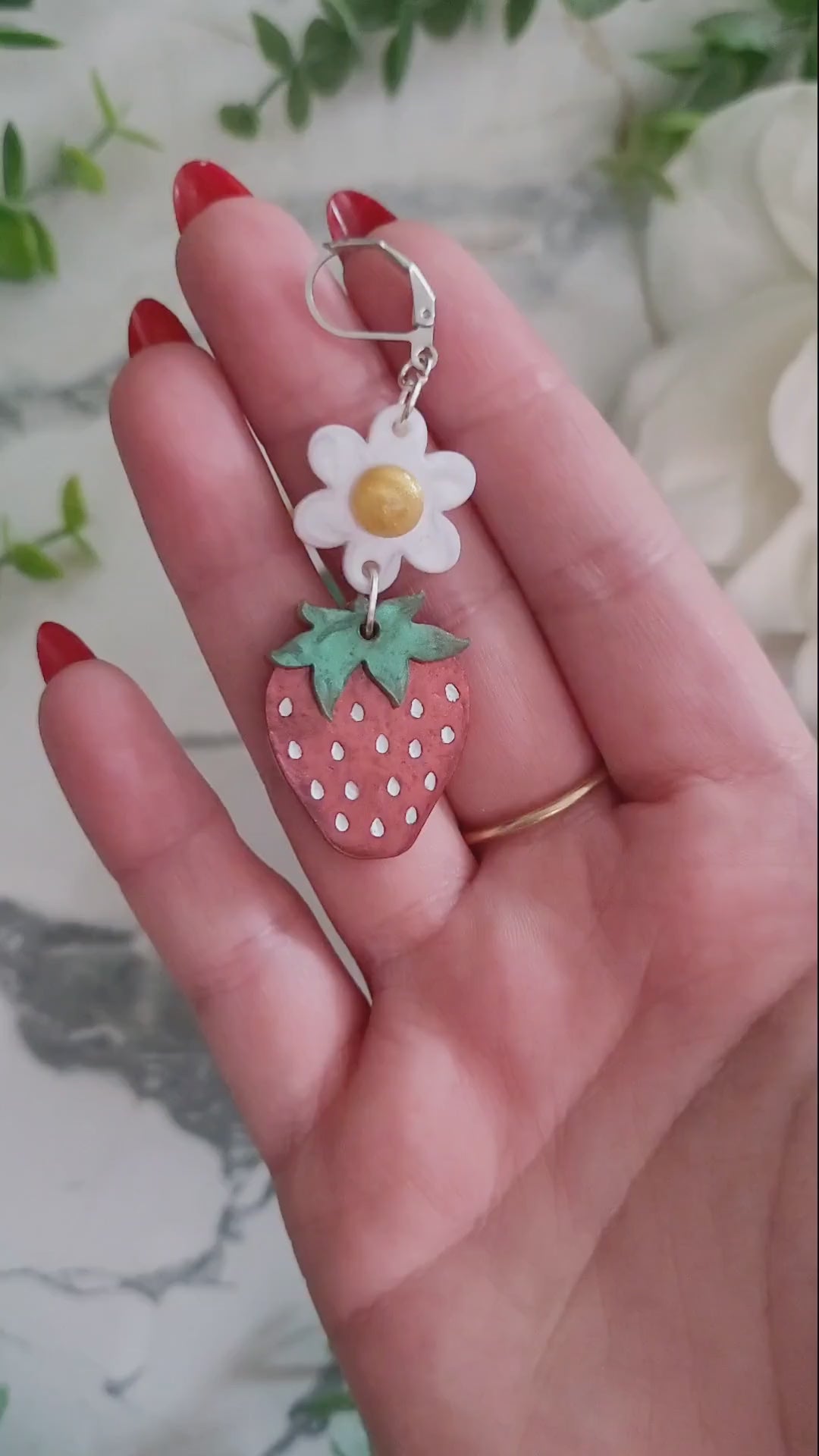 video close up of Resin Strawberry and flower earrings with gold and silver findings on a marble background surrounded by foliage.
