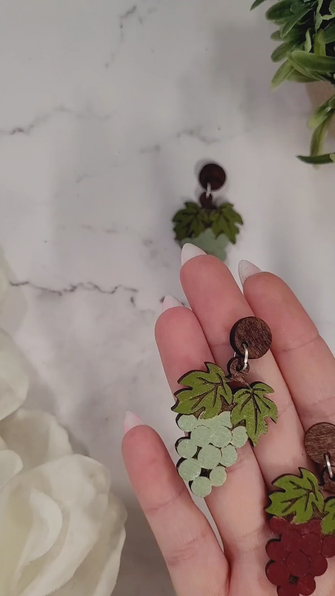 video close up of  red and Green grape bunch earrings on a marble background surrounded by foliage