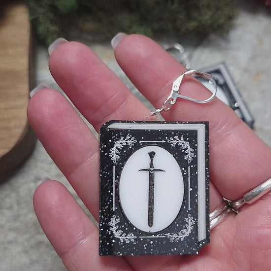 video of Glitter sword Book Earrings on a stone background surrounded by foliage. 
