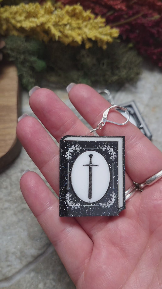 video of Glitter sword Book Earrings on a stone background surrounded by foliage. 