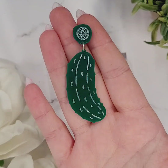 video close up of Green pickle earrings on a white marble background surrounded by foliage.