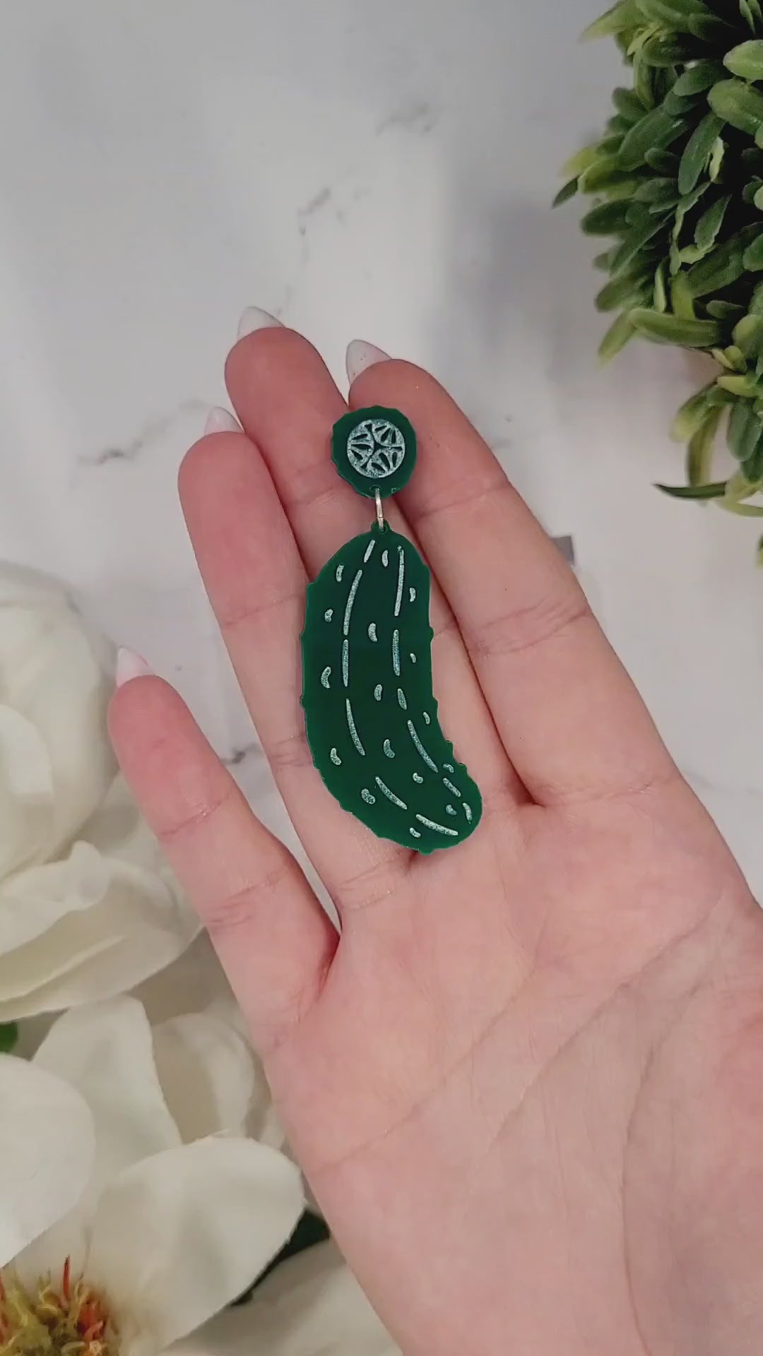 video close up of Green pickle earrings on a white marble background surrounded by foliage.