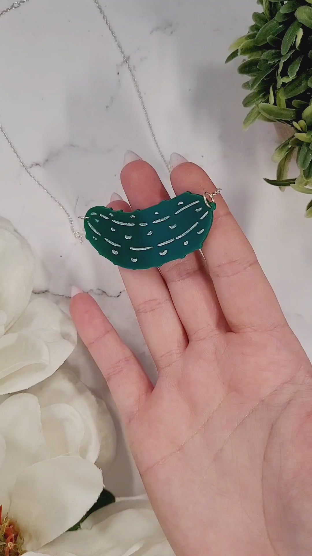 video close up of Green pickle necklace on a white marble background surrounded by foliage.