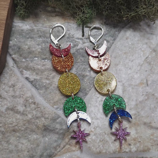 video of Rainbow moon phase earrings on a stone background surrounded by foliage 