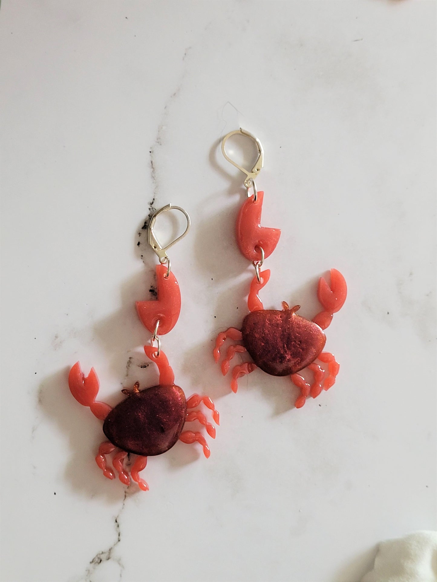 Close up of resin hook earrings in the shape of a red crab with metallic accents. 