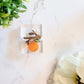 Resin brooch of tree branch with a flower, orange, and leaf dangling from it. 
